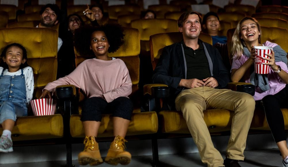 Grups de persones adultes i nens asseguts en butaques grogues d'una sala de cinema rient i menjant crispetes