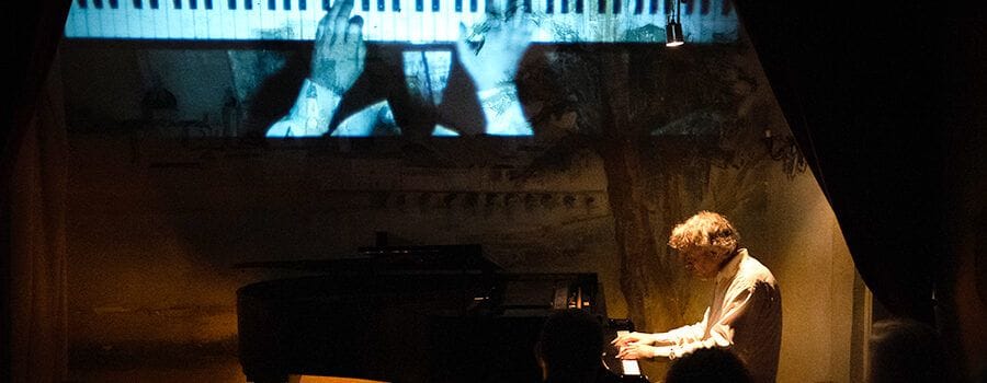 Pianista al piano en un escenari fosc, amb una projecció en blanc i negre de mans sobre tecles al fons.