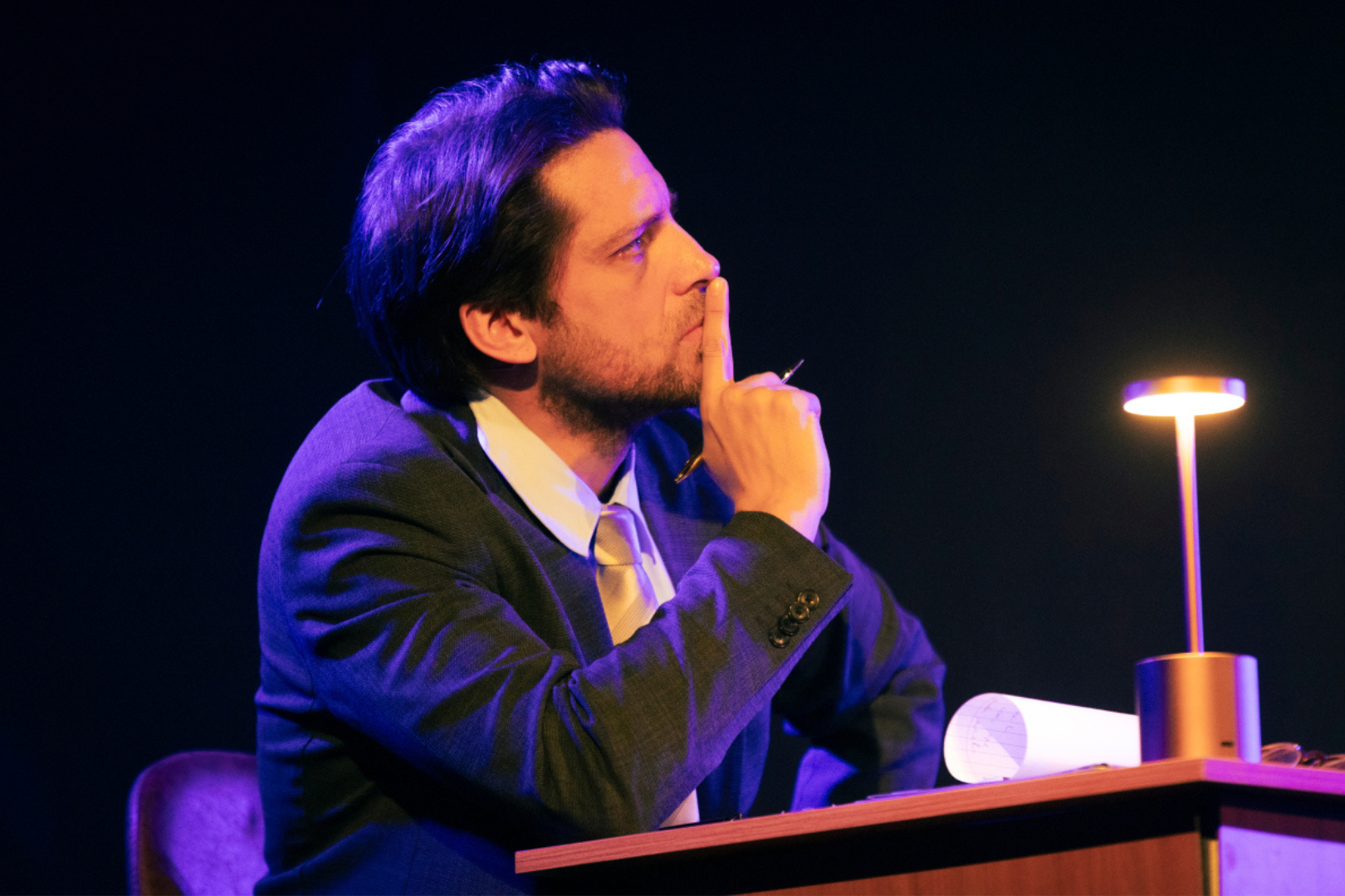 Home amb vestit assegut a una taula a l'escenari fosc, pensatiu, il·luminat per un llum de taula