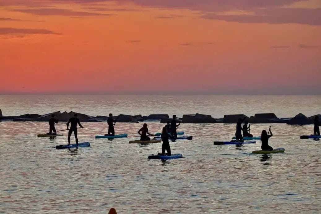 Siluetes de persones dempeus sobre paddleboards al mar en calma amb un cel taronja intens i esculls rocosos al fons
