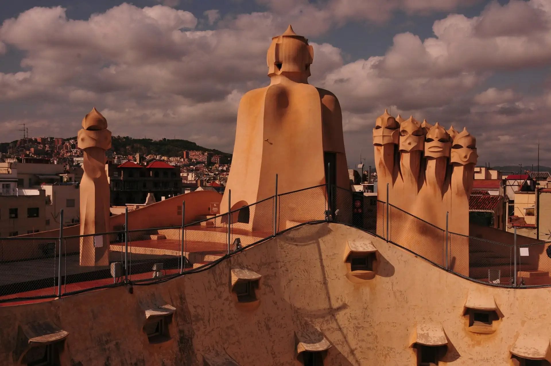Vista panoràmica des de la terrassa d'un edifici modernista amb xemeneies esculturals i un ampli paisatge urbà al capvespre.
