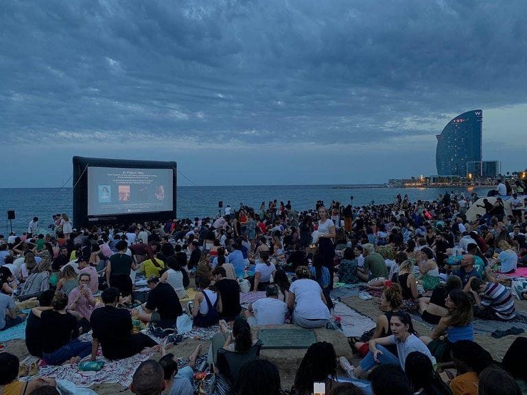 Multitud asseguda a la sorra d'una platja al capvespre gaudint d'una projecció en pantalla gegant a l'aire lliure