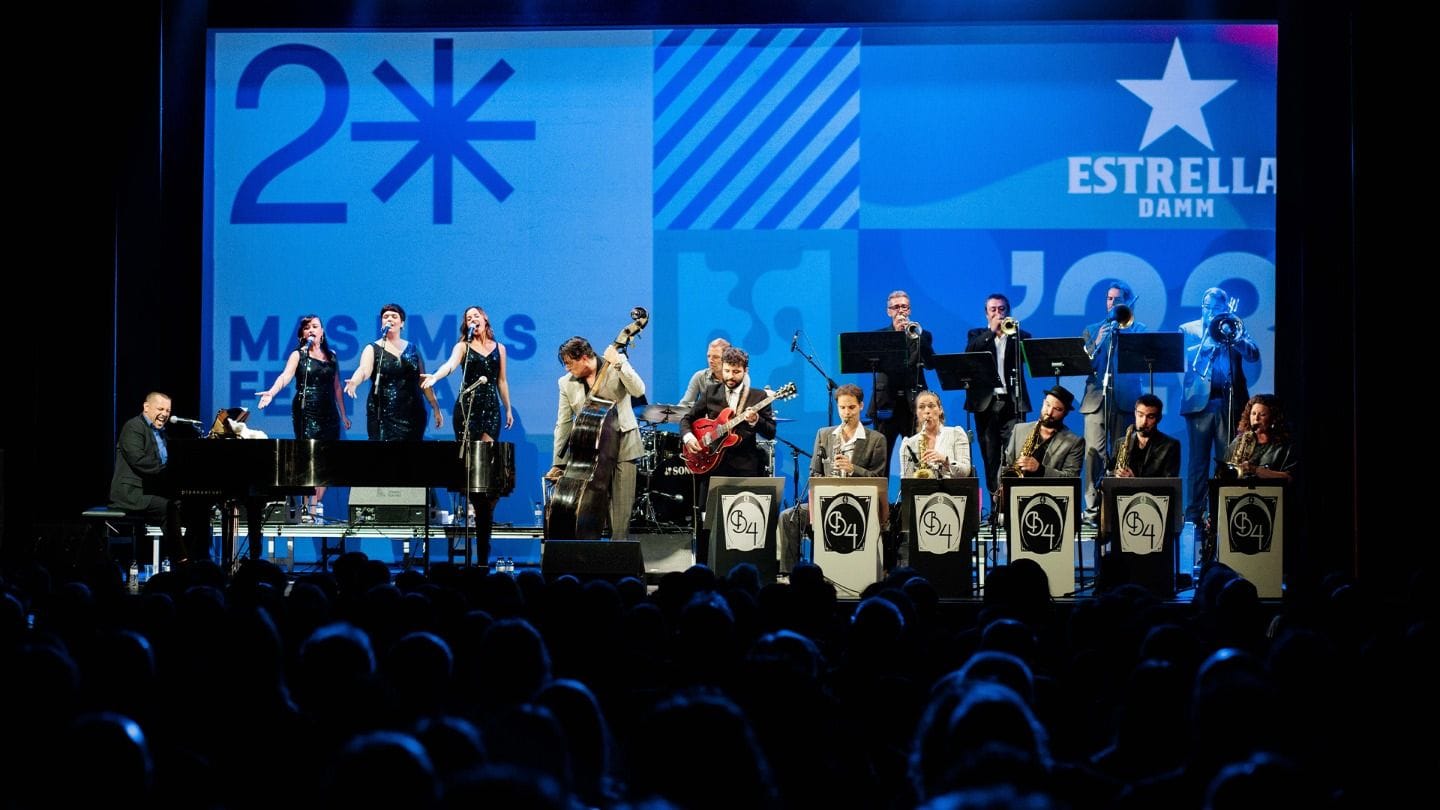 Gran orquestra sobre un escenari davant d'un gran plafó blau, amb pianista al capdavant i públic en silueta.