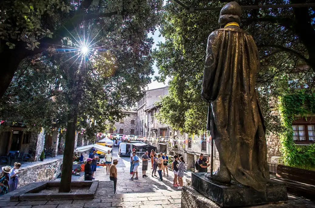 Estàtua de pedra d'una figura amb robes llargues en primer pla, amb una plaça assolellada amb arbres i vianants al fons.
