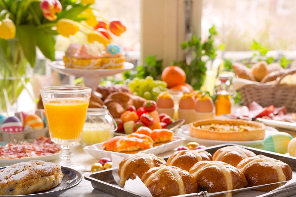 Taula de brunch amb panets creuats, suc de taronja, pastes, fruita i flors grogues vora la finestra