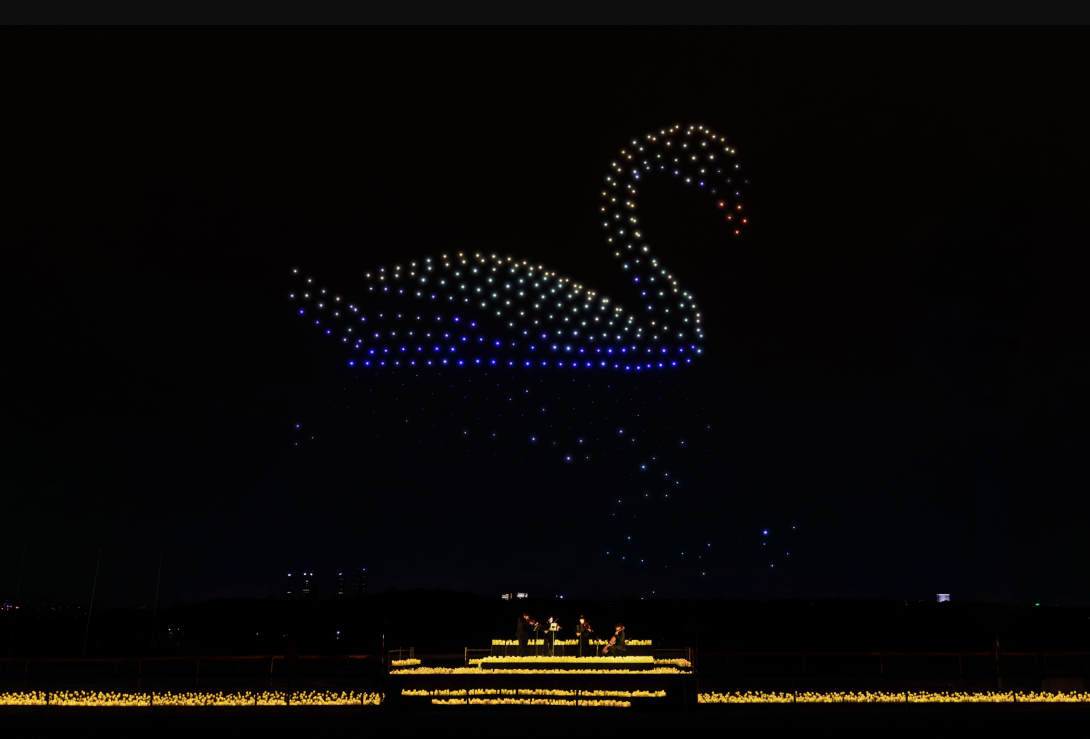 Centenars de drons il·luminats formen la silueta d'un cigne blanc al cel nocturn sobre un escenari flotant al riu.