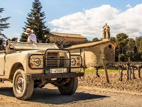 Vehicle tot terreny vintage aparcat enmig d'una vinya amb una capella de pedra al fons i ceps nus en primer pla.