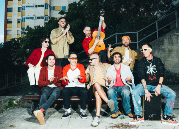 Grup d'unes deu persones amb roba de colors posen a l'exterior; un d'ells sosté una guitarra acústica