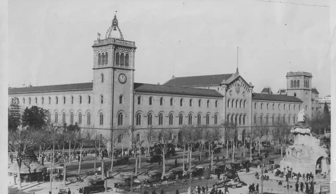 Fotografia històrica en blanc i negre d'un gran edifici neoclàssic amb torre de rellotge, davant d'una avinguda amb vehicles i vianants d'època.
