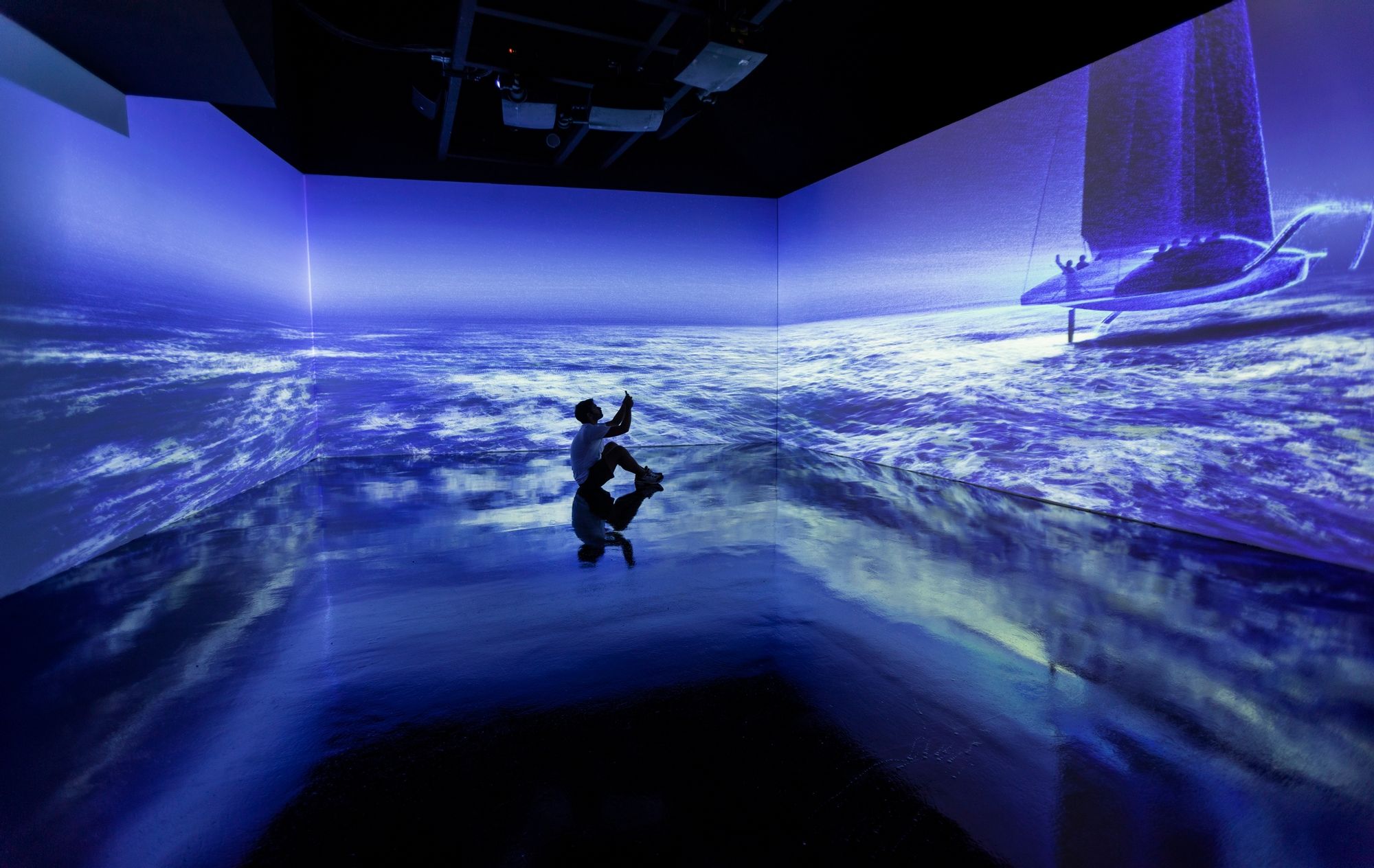 Sala immersiva fosca amb projeccions d'onades oceàniques en blau i la silueta d'una vela al fons