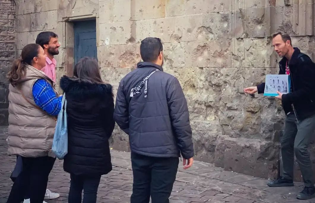 Grup de persones al carrer de pedra escoltant un guia que sosté documents, tots amb jaquetes d'hivern.