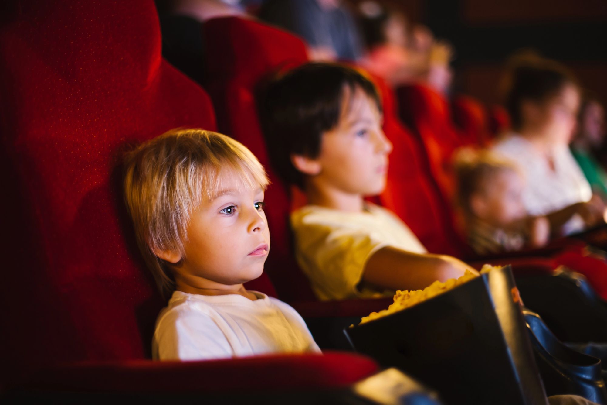 Nens petits asseguts en butaques vermelles de cinema mirant una pel·lícula, un d'ells amb crispetes a les mans