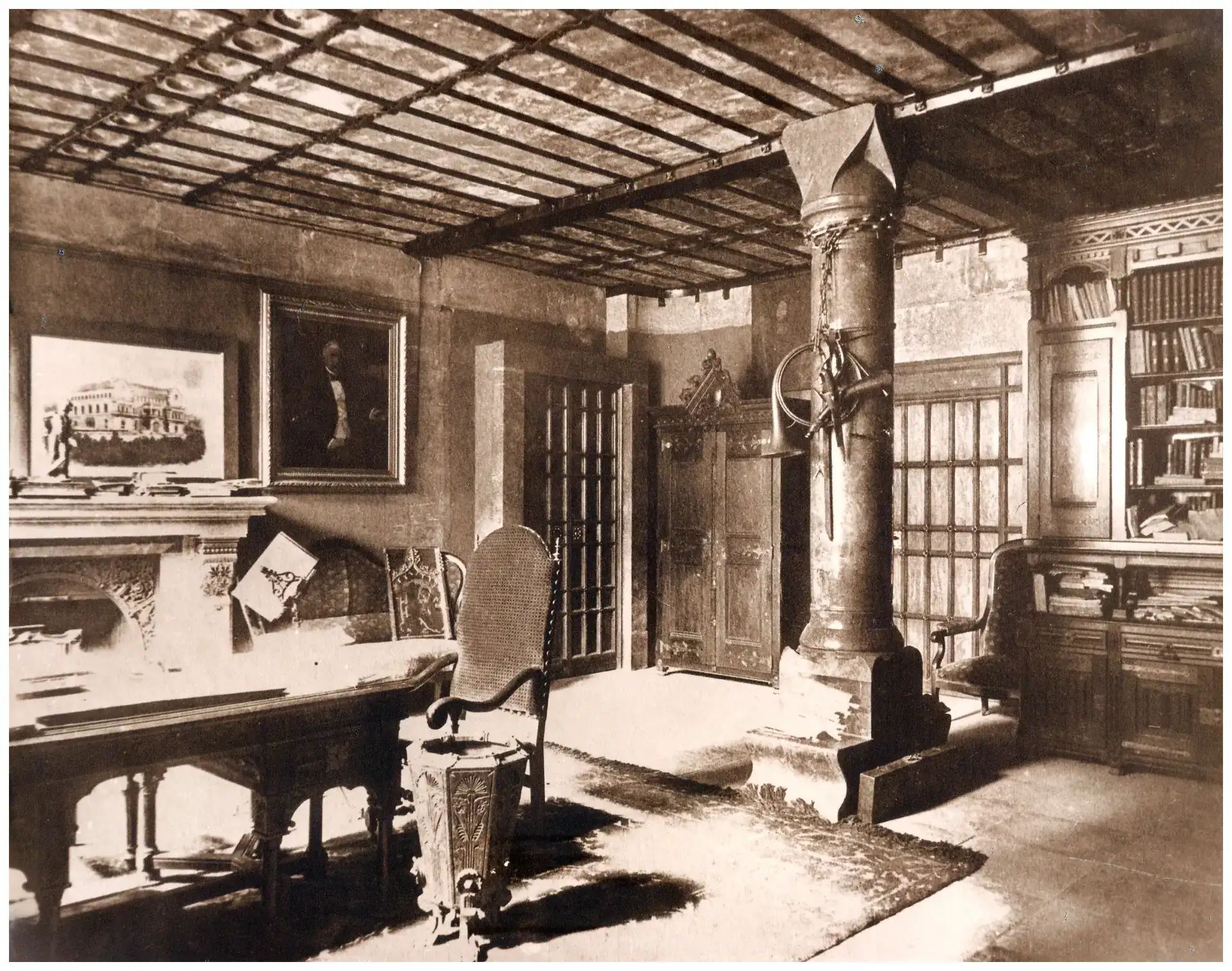 Fotografia històrica en blanc i negre d'una sala interior amb columna de fusta ornamentada, llar de foc i mobiliari antic.