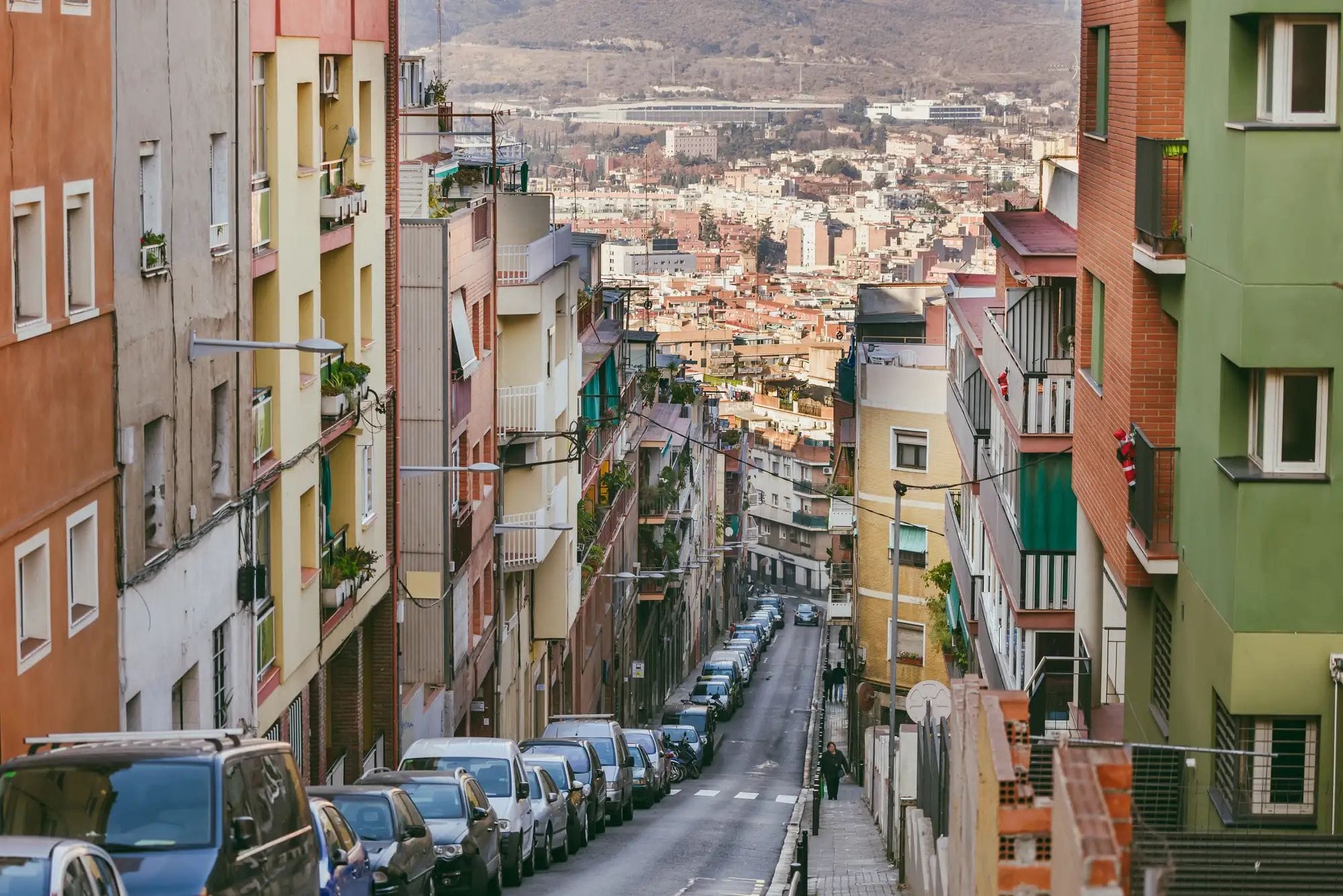 Carrer urbà en fort pendent flanquejat per edificis d'apartaments de colors i cotxes aparcats, amb la trama de la ciutat al fons.