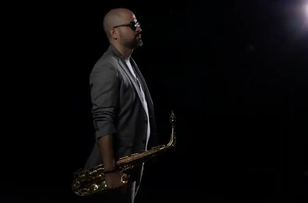 Músic de perfil amb blazer gris sostenint un saxofon sobre fons negre