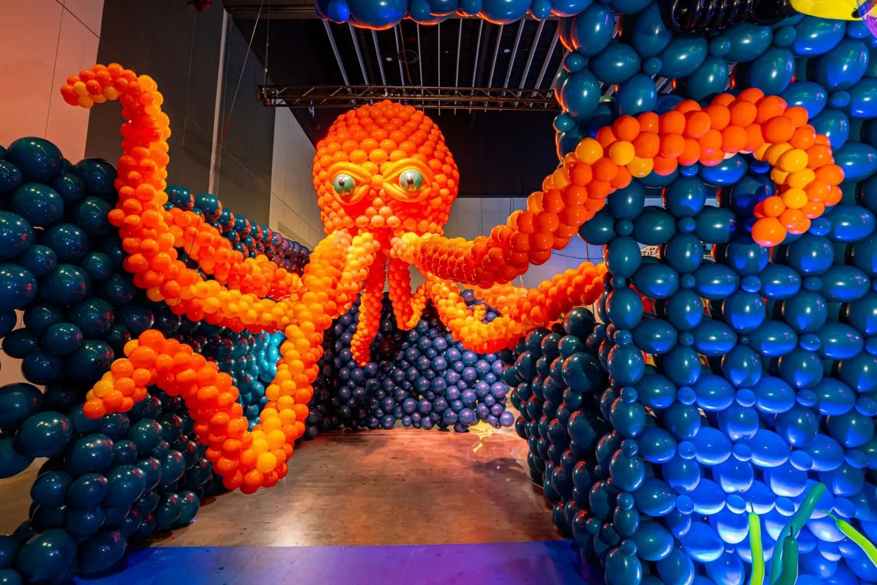 Escultura gegant de pop feta amb globus taronges, amb tentacles estesos, en un espai interior fosc amb parets de globus blaus.