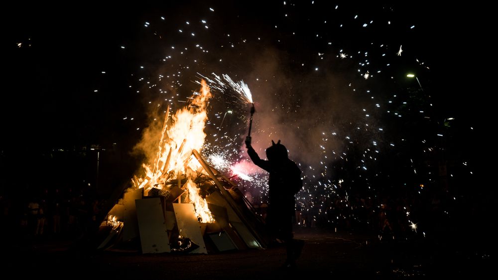 Silueta d'una persona amb una torxa encesa al costat d'una gran foguera amb espurnes al cel nocturn