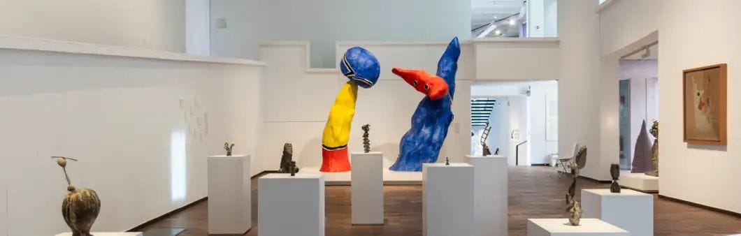 Sala de museu lluminosa amb escultures abstractes de gran format en blau, vermell i groc sobre pedesals blancs, envoltades de peces més petites.